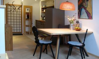 A cozy dining area with a wooden table and two chairs. In the background, there is a modern kitchen and a colorful picture on the wall.