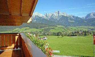 A beautiful landscape with mountains in the background and green meadows. On a balcony, there are flowers, and the sky is clear and blue.