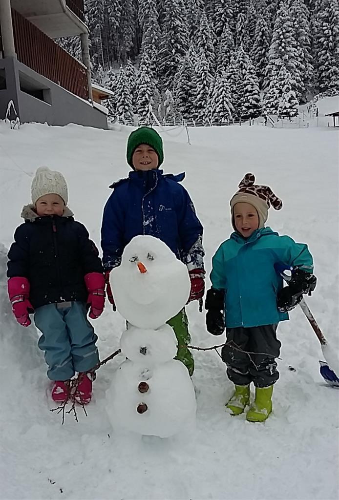 Three children are standing in the snow and smiling next to a snowman. The snowman has a carrot nose and eyes made of coal.