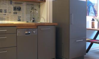 A modern kitchen with gray cabinets and a wooden countertop. In the background, dishes and glasses can be seen on a shelf.