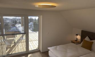 A bright bedroom with a comfortable double bed and a window that opens to a balcony with a view. The warm lighting creates an inviting atmosphere.