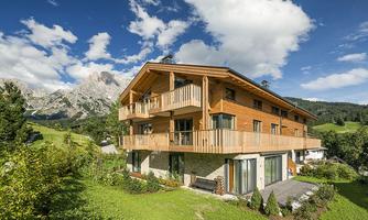 A modern wooden house in a picturesque alpine landscape. The sky is blue, and mountains can be seen in the background.