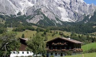 A picturesque mountain landscape with high, snow-capped peaks. In the foreground stands a traditional house with balconies and a green meadow.
