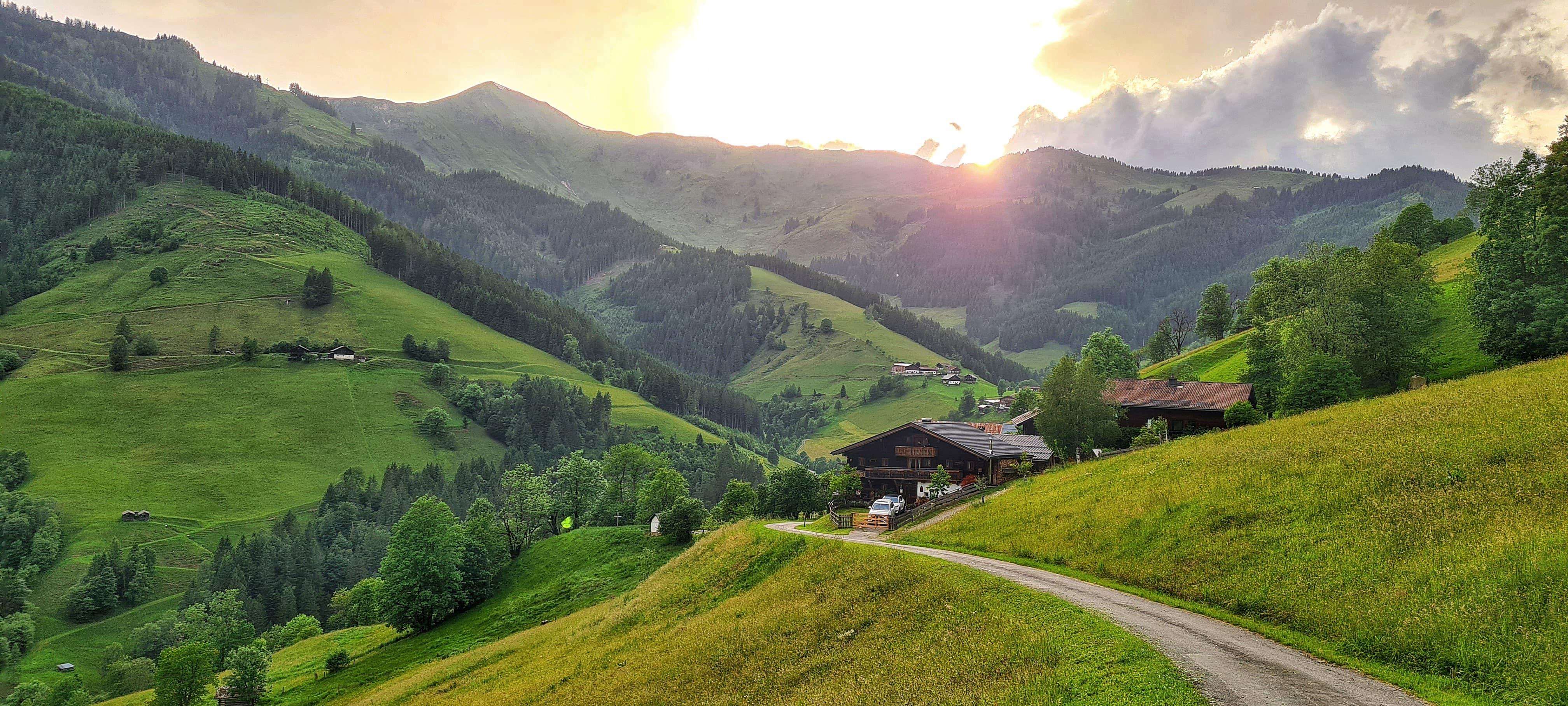 A picturesque landscape with gentle hills and green meadows. The sun sets behind the mountains, giving the scene a warm light.