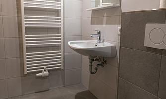 A modern bathroom with gray tiles and a white sink. A towel warmer is hanging on the wall and a bath towel is lying on the toilet.