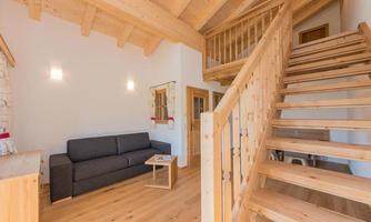 A cozy living room with a gray sofa and a wooden table. A bright staircase leads upstairs, surrounded by wood paneling.