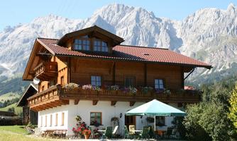A charming wooden house with a balcony and colorful flowers. Majestic mountains rise in the background under a clear sky.
