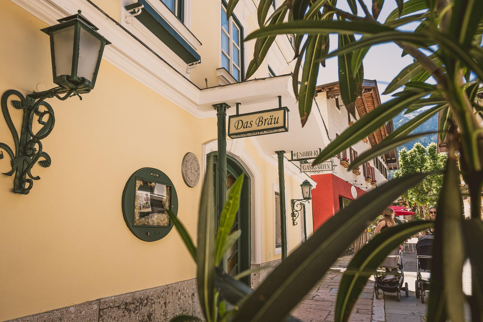 A nice inn called "Das Bräu" can be seen on a colorful street. Next to the entrance door, there are plants and a wall lamp.