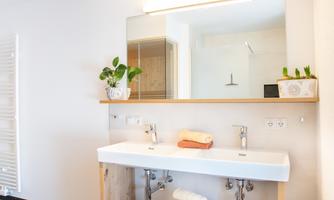 A modern bathroom with a double sink and wooden shelf. Plants and towels create an inviting atmosphere.