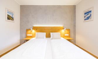 A modern bedroom with a double bed and soft wooden tones. Pictures hang on the wall and warm white lighting creates a cozy atmosphere.