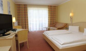 A bright, cozy hotel room with a double bed and a seating area. There is a desk, a television, and large windows with curtains.