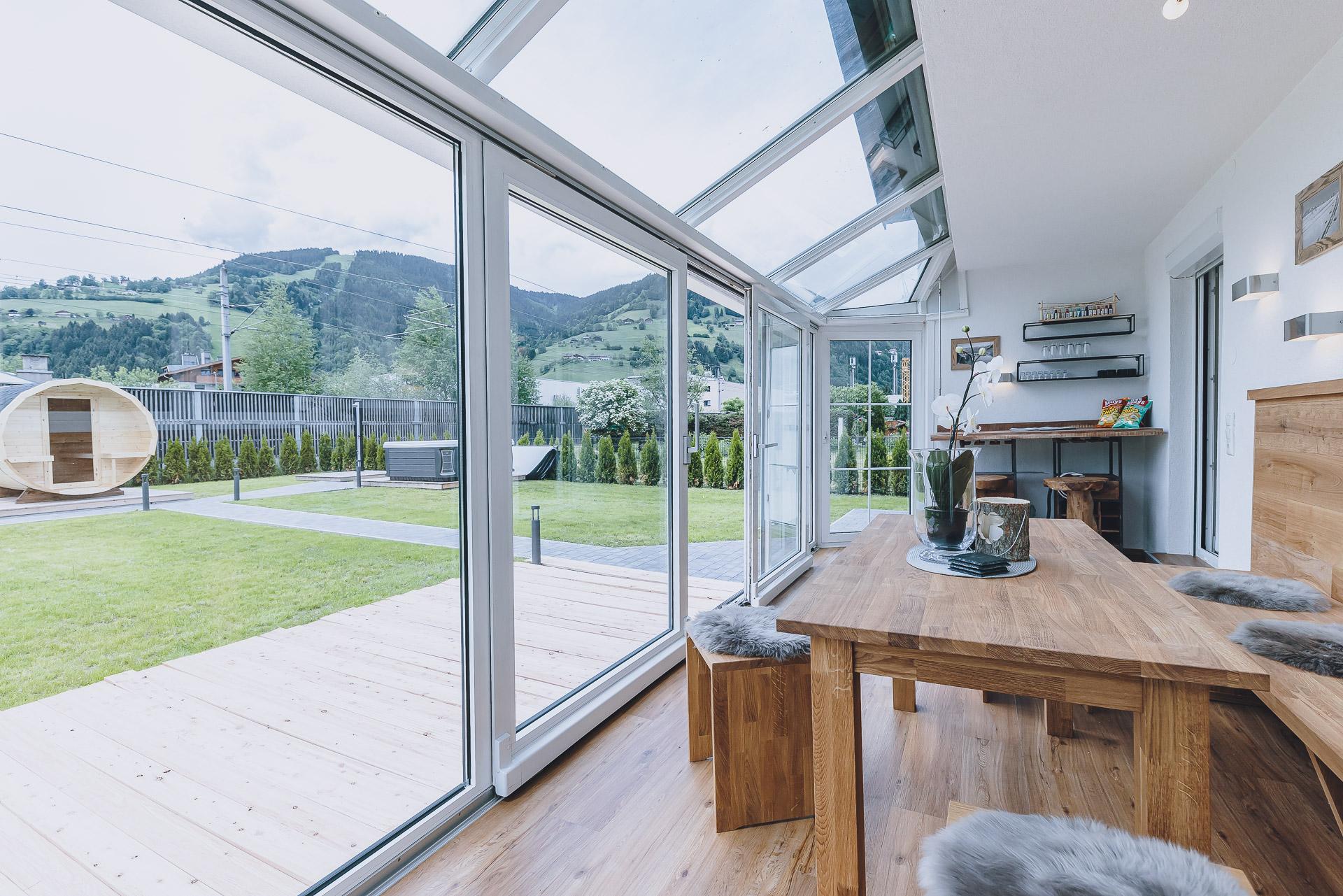 A bright conservatory with large windows and a wooden table. In the background, green meadows and hills stretch out.