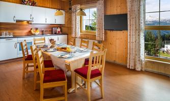 An inviting dining area with a large table and wooden chairs. The kitchen is modern and well-equipped, overlooking the nature outside.