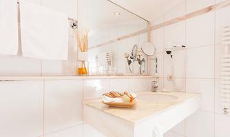 A modern bathroom with white tiles and a sink. On the shelf, there are towels and a decorative container with skincare products.