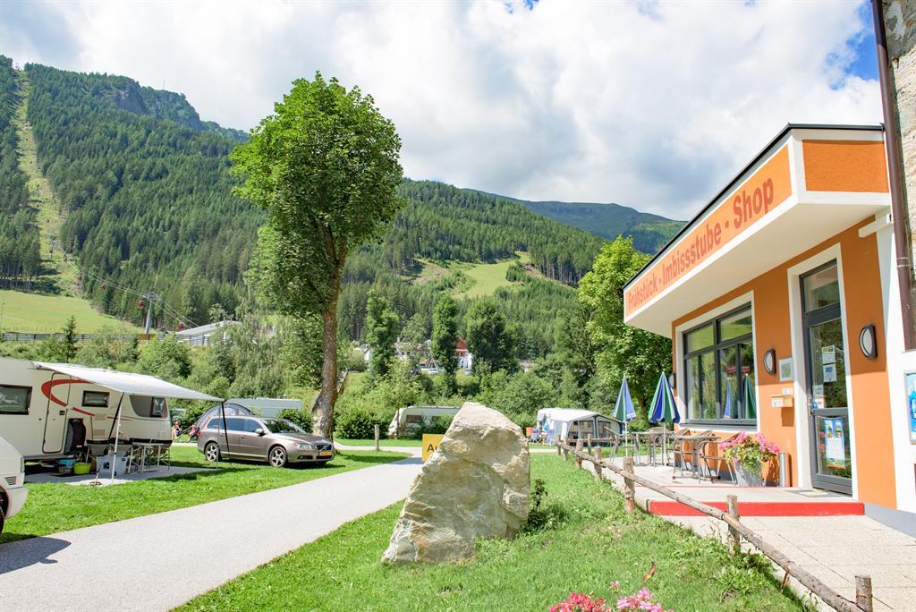 A campsite in the mountains with motorhomes and a kiosk. The surroundings are green and it is sunny.