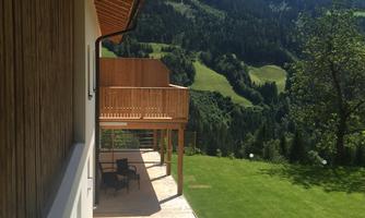A modern house with a wooden veranda and a view of green hills. The sky is blue and the landscape is lush and mountainous.