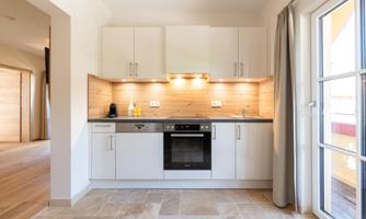 A modern kitchen with white cabinets and wooden details. It features an oven, a sink, and a window that allows for plenty of daylight.