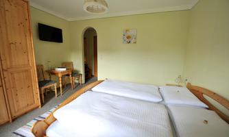 A simple bedroom with two single beds and a TV on the wall. There is a small table with chairs and a wooden wardrobe.