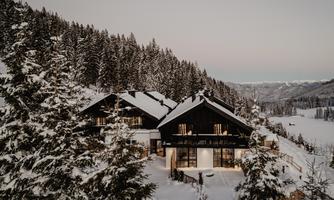 A modern house in a snowy environment, surrounded by tall fir trees. The landscape is calm and wintry, with a gentle sky in the background.