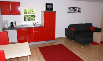 A modern apartment with a red kitchen unit and a cozy black-gray sofa. The room features a wooden floor and a red carpet.