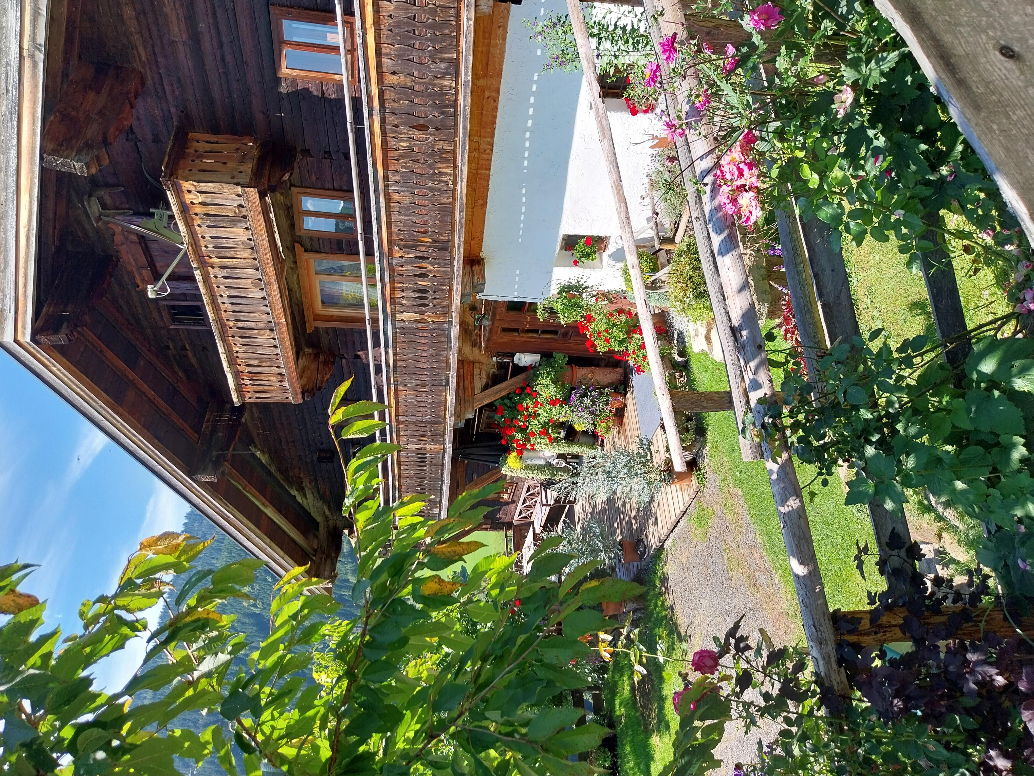 A traditional wooden house with a balcony and blooming plants in the foreground. The surroundings are green and sunny.