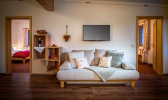 A cozy living space with a bright sofa and decorative cushions. In the background, there is a bedroom and a television on the wall.