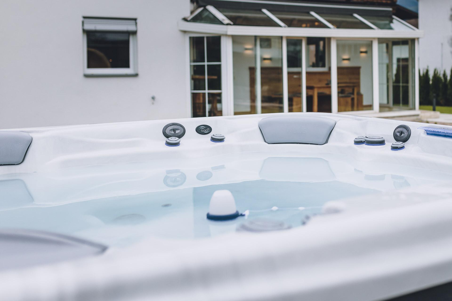 A modern outdoor hot tub, with clear water and comfortable seating. In the background, there is a bright, welcoming home.