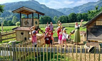 A group of happy children stands in a garden with a small stable and meadows in the background. The children are waving and having fun in nature.