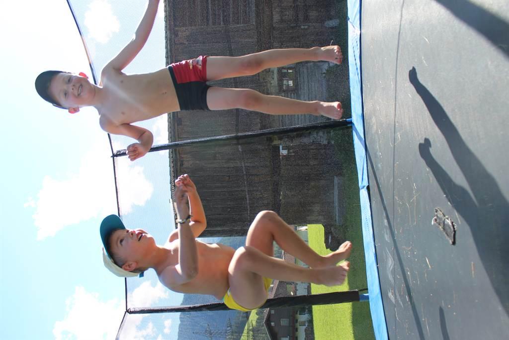 Two children are happily jumping on a trampoline. The sky is blue and the sun is shining.