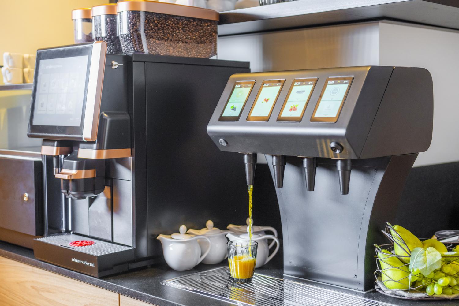 A modern coffee machine stands next to a drink dispenser. Both devices are in a bright kitchen and ready to prepare various drinks.