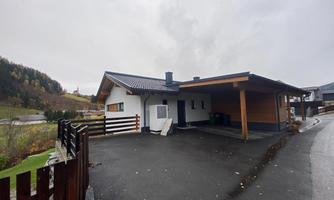 A modern house with a covered carport. The surroundings are green and mountainous, it is cloudy.
