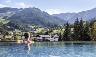 A person relaxes in an infinity pool overlooking green hills and mountains. The landscape is calm and picturesque, under a clear blue sky.