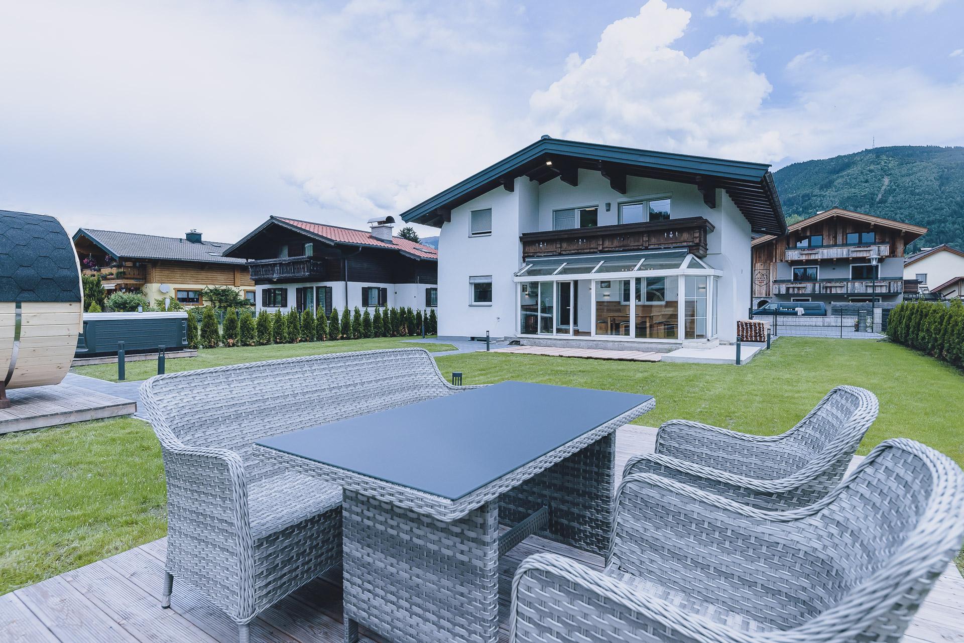 A modern house with a well-kept garden and rattan terrace furniture. In the background, the mountains and other buildings can be seen.