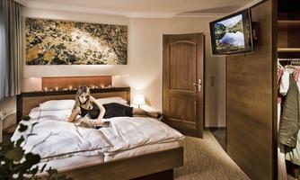 A modern bedroom with a large bed and a wooden closet. A woman is sitting on the bed reading, while a television is playing in the background.