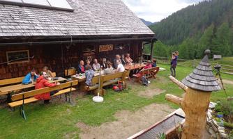 A group of people is sitting at tables in front of a rustic cabin in nature. In the background, green hills and a cloudy sky can be seen.
