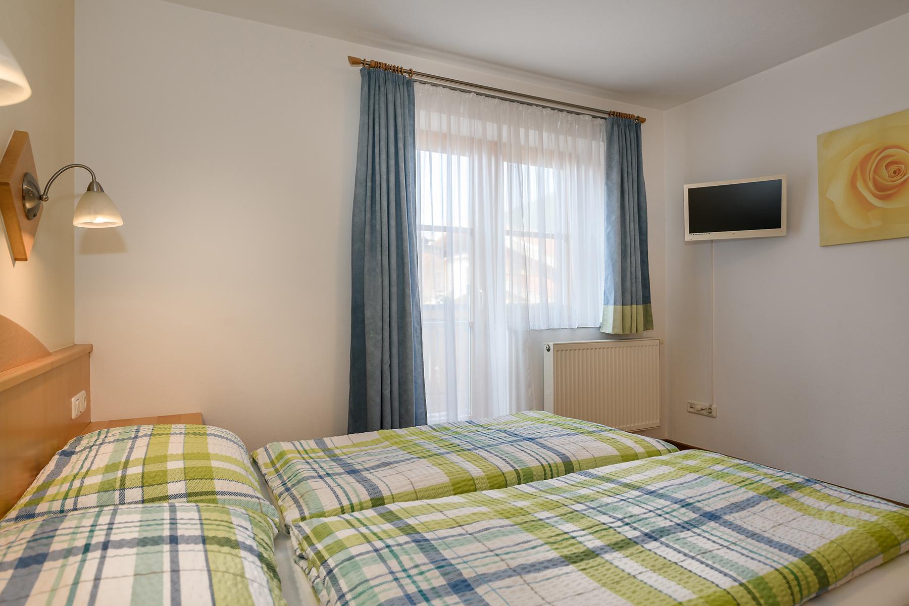 A bedroom with a double bed and checkered bedspreads. A window with curtains and a television on the wall, as well as a large wardrobe, are also visible. Quiet and central location in Dorfgastein. The mountain lifts, ski lifts, and swimming pool are just a few meters away. Good restaurants nearby.