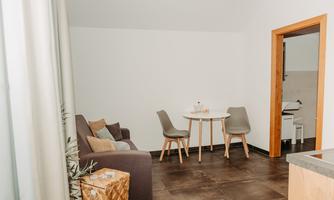 A modern living room with a gray sofa, a small table, and chairs. The room is bright and simply designed, with a view of an open door to the bathroom.