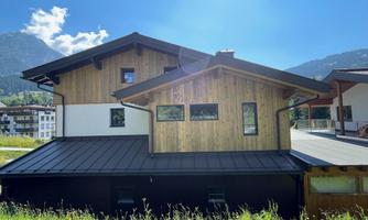 A modern wooden house with a flat roof. In the background, mountains and a blue sky can be seen.