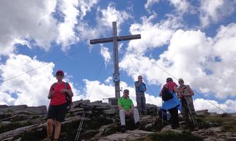A group of hikers stands at a summit cross in nature. The sky is blue with some clouds.