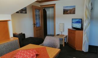 A cozy living room with a dining table and comfortable seating. Pictures hang on the wall, and a television stands on a wooden cabinet.