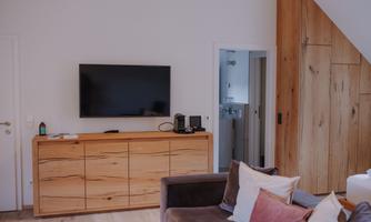A modern living space with a television on the wall and a wooden cabinet. In the foreground, there is a cozy sofa with colorful cushions.