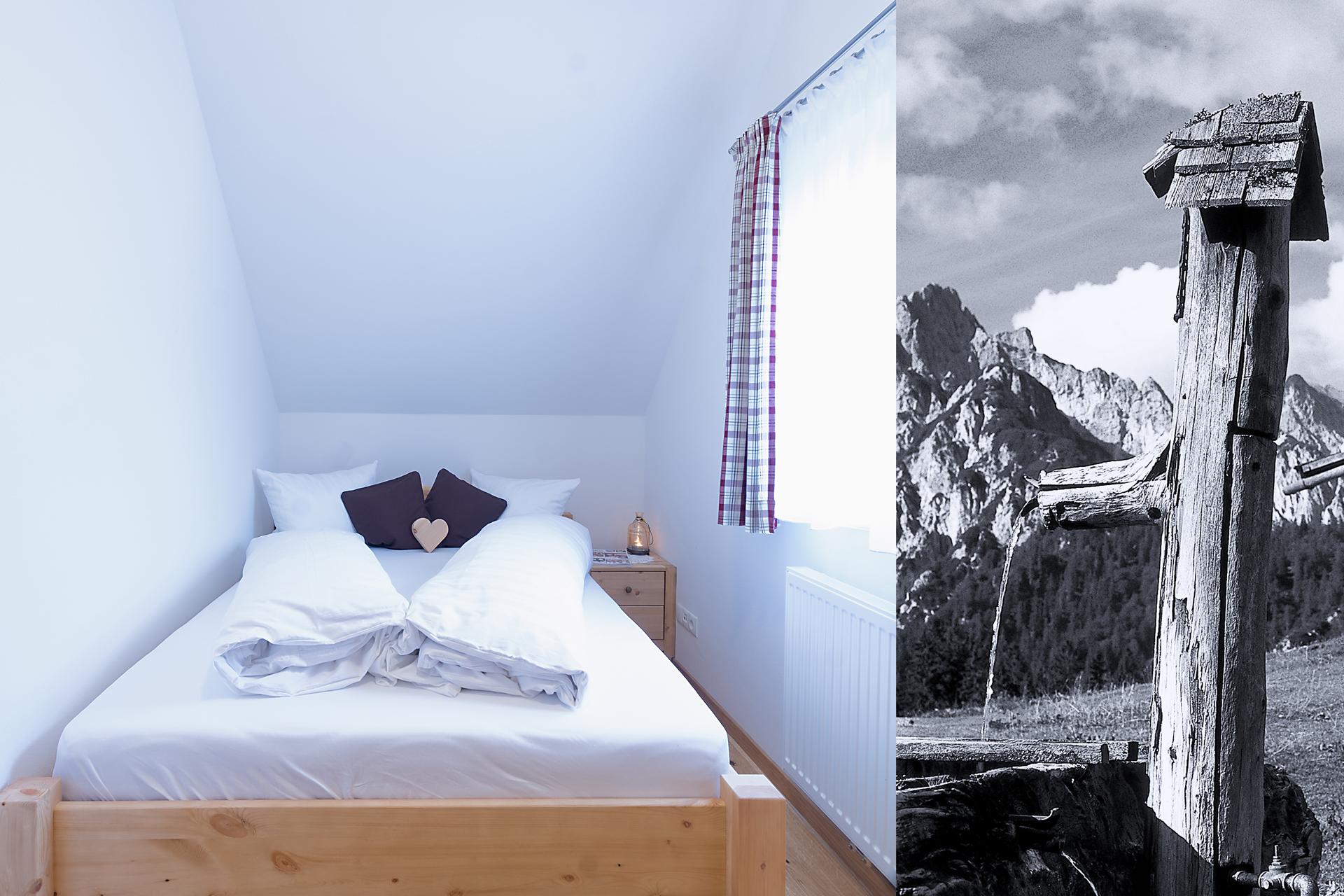 A comfortable bedroom with a wooden bed and a window with curtains. On the other side, a traditional well is visible in a mountain landscape.