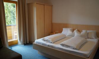 A modern bedroom with a large double bed and fresh white bedding. The room has a balcony and bright curtains.
