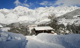 A snowy winter scene with majestic mountains in the background. In the foreground is a path leading to a wooden house, surrounded by fresh, white snow.