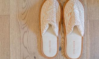 Comfortable house slippers with a modern, minimalist design are placed on a wooden floor. The shoes are light and have a fine texture.
