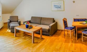 A cozy living space with a sofa, a table, and chairs. The bright atmosphere is enhanced by large windows and a wooden floor.