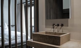 A modern bathroom with a minimalist sink and a large mirror. In the background, a bedroom with a large window and curtains is visible.