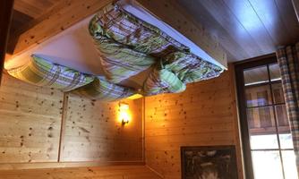 A cozy room with wooden paneling and a double bed. The ceiling is decorated with colorful cushions and a window lets in daylight.