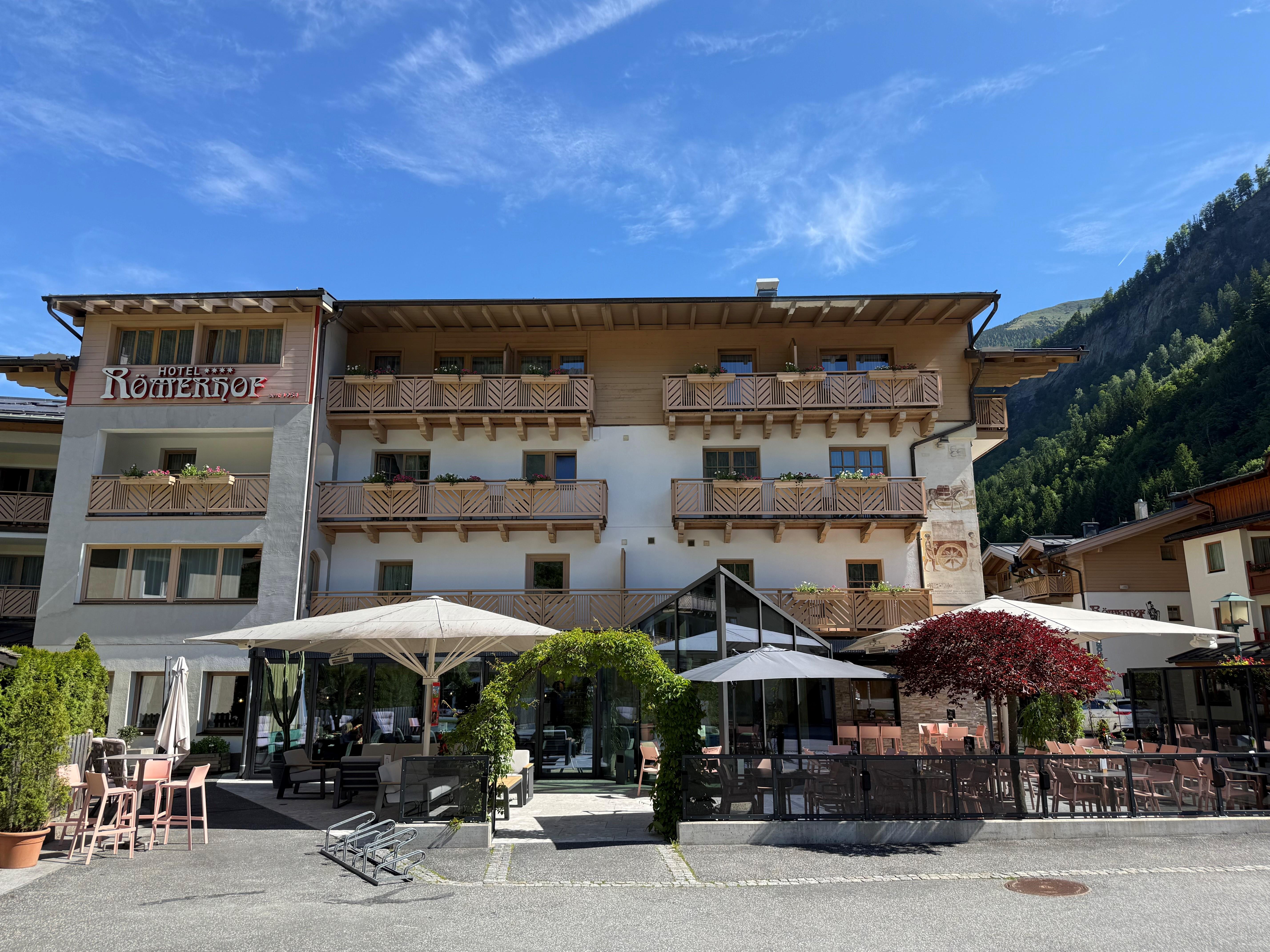 A beautiful hotel with several balconies and an inviting terrace. The clear sky and the surrounding mountains create a pleasant atmosphere.