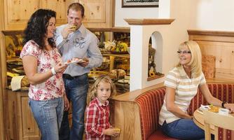 A family is enjoying their meal in a cozy room with wooden decor. In the background, food and wall decoration can be seen.